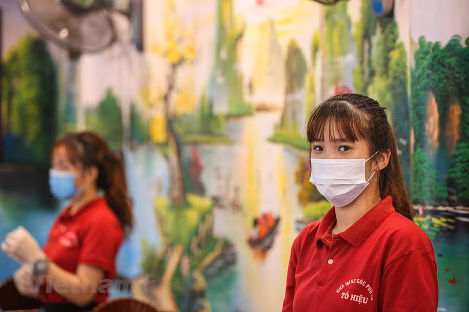 Todos los empleados en los restaurantes deben usar mascarillas para atender a los clientes e implementar drásticamente las medidas destinadas a prevenir la propagación epidémica de COVID-19. La Organización Mundial de la Salud (OMS) la reconoció como una pandemia global el 11 de marzo de 2020. El coronavirus se transmite generalmente de persona a persona a través de las pequeñas gotas de saliva, conocidas como microgotas de Flügge, que se emiten al hablar, estornudar, toser o espirar. Se difunde principalmente cuando las personas están en contacto cercano, pero también se puede trasmitir al tocar una superficie contaminada y luego llevar las manos contaminadas a la cara o partes del cuerpo con mucosa. Su período de incubación suele ser de cinco días, pero puede variar de dos a 14 días. (Foto: Minh Son / Vietnam +)
