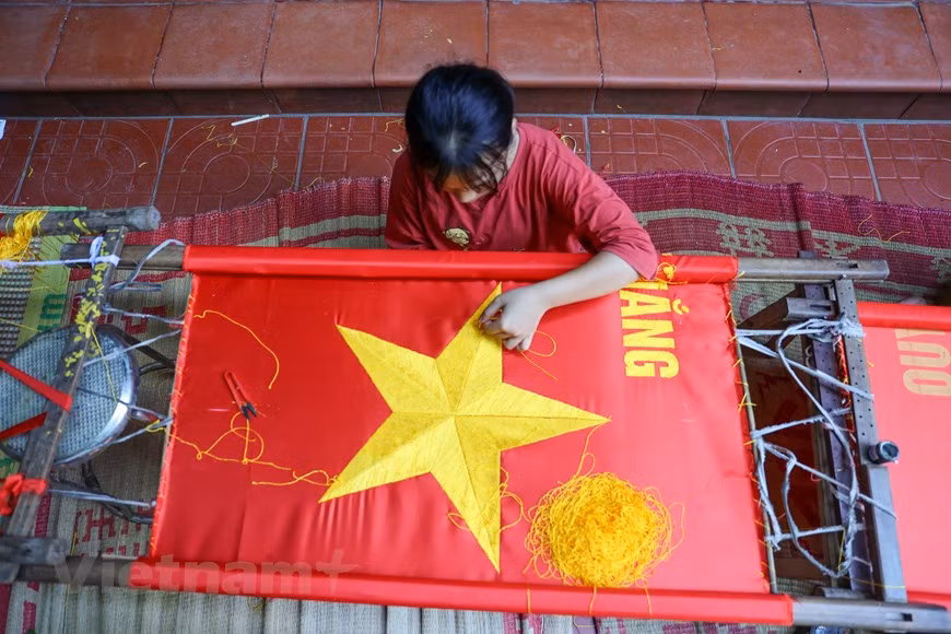 Para poder producir una bandera hecha a mano, la tela utilizada debe proceder de la aldea de La Khe, en el distrito de Ha Dong, en Hanoi. La pieza de tejido debe poseer una textura gruesa y un color rojo brillante, para brindar durabilidad y resistencia ante las condiciones climáticas. Vuong Thi Nhung reveló que existen pedidos de hasta miles de banderas de grandes tamaños, que se utilizarán principalmente para los desfiles militares. También dijo que coser banderas con máquinas es más simple que bordarlas. Una máquina puede coser cientos de banderas en un día, pero el bordado se demora hasta semanas. (Foto: Vietnam+)