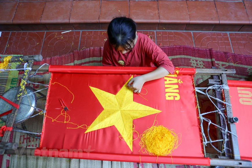 Para poder producir una bandera hecha a mano, la tela utilizada debe proceder de la aldea de La Khe, en el distrito de Ha Dong, en Hanoi. La pieza de tejido debe poseer una textura gruesa y un color rojo brillante, para brindar durabilidad y resistencia ante las condiciones climáticas. Vuong Thi Nhung reveló que existen pedidos de hasta miles de banderas de grandes tamaños, que se utilizarán principalmente para los desfiles militares. También dijo que coser banderas con máquinas es más simple que bordarlas. Una máquina puede coser cientos de banderas en un día, pero el bordado se demora hasta semanas. (Foto: Vietnam+)