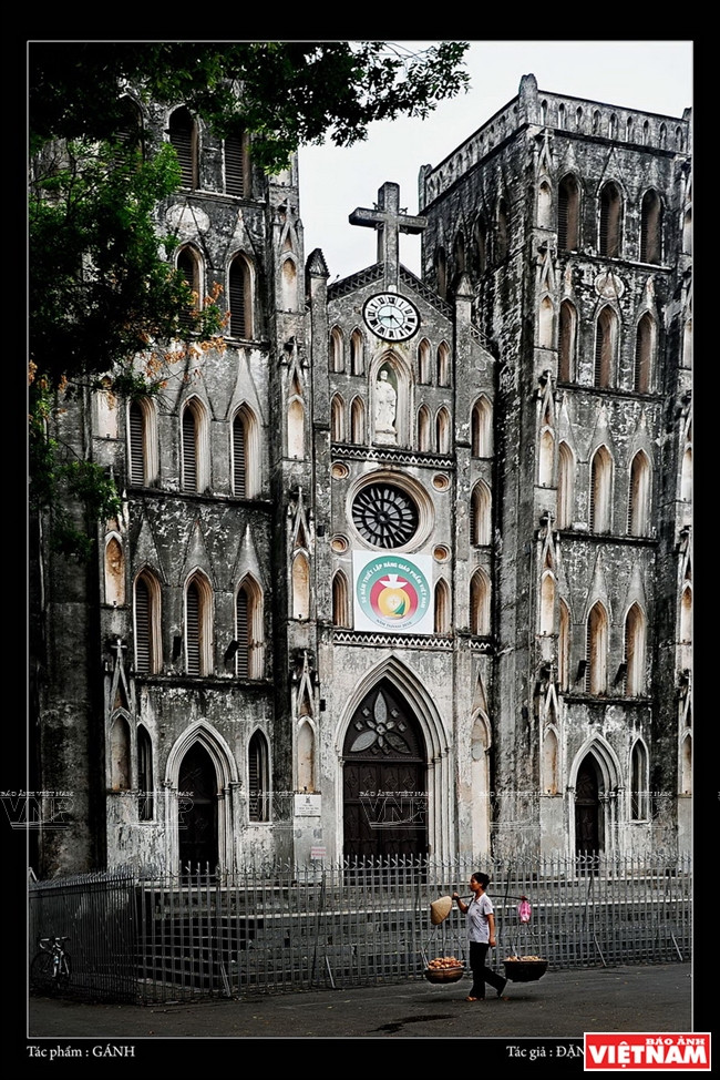 “Carga” – una mujer llevando en los hombros la supervivencia de su familia camina en frente de la Catedral de Hanoi.